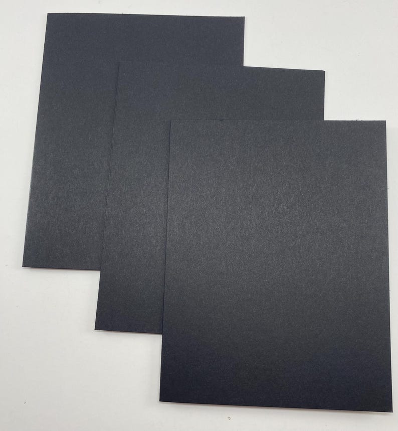 Pode incluir: Uma pilha de tr&ecirc;s folhas de papel cart&atilde;o preto. O papel tem uma superf&iacute;cie ligeiramente texturizada e &eacute; retangular. As folhas est&atilde;o dispostas em um padr&atilde;o escalonado, com cada folha sobrepondo-se parcialmente &agrave; de baixo.