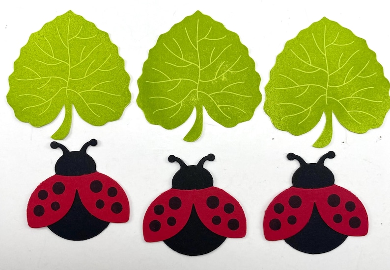 Pode incluir: Tr&ecirc;s recortes de folhas verdes e tr&ecirc;s recortes de joaninhas. As joaninhas t&ecirc;m asas vermelhas com pintas pretas e corpos pretos. As folhas s&atilde;o verde claro com padr&otilde;es de veias detalhados.
