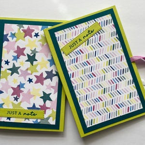 Puede incluir: Dos cuadernos hechos a mano con cubiertas decorativas. Uno presenta un patrón de estrellas en rosa, verde y amarillo. El otro tiene un diseño de rayas diagonales coloridas. Ambos tienen un borde verde, un marco amarillo y el texto "JUST A note".
