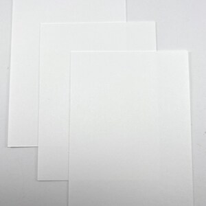 Pode incluir: Tr&ecirc;s folhas de papel retangulares brancas, ligeiramente sobrepostas. O papel tem um acabamento liso e fosco e est&aacute; disposto sobre um fundo branco liso. As folhas variam ligeiramente de tamanho, criando um efeito em camadas.