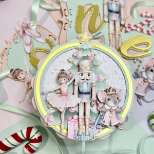 Op de afbeelding: Een grillige kersttaarttopper met een notenkraker, ballerina en muis met een kroon. Het ontwerp bevat een kerstboom, cadeaus en de letter "n" in een feestelijk lettertype. Het kleurenpalet bevat roze, blauwe en gouden accenten.