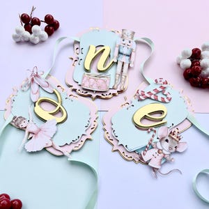 May include: A decorative banner spelling "one" with gold letters, featuring a ballerina, a nutcracker, and a mouse. The banner is in pastel pink and blue with gold accents, ideal for a celebration. Berries are in the background.