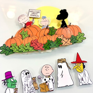 May include: A Peanuts-themed decorative set featuring a pumpkin patch with Linus, Sally, and Snoopy, plus a sign reading WELCOME GREAT PUMPKIN. Below are five individual character cutouts including figures in ghost costumes and a witch, all in vibrant colors.