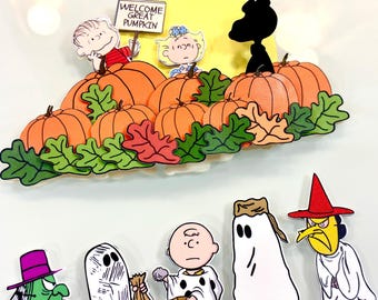 May include: A Peanuts-themed decorative set featuring a pumpkin patch with Linus, Sally, and Snoopy, plus a sign reading WELCOME GREAT PUMPKIN. Below are five individual character cutouts including figures in ghost costumes and a witch, all in vibrant colours.