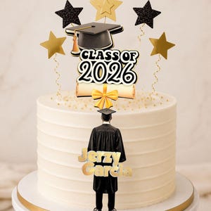 Puede incluir: Un adorno para tarta de graduación con un birrete negro, un diploma y el texto "CLASS OF 2026" en blanco y negro. Incluye estrellas doradas y negras con purpurina en tallos de alambre y una figura de un graduado con el nombre "Jerzy Garcia".