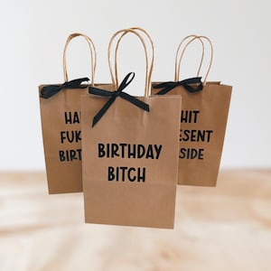 May include: Three brown paper gift bags with black text and black ribbon bows. The bags have the phrases "HA FUK BIRT", "BIRTHDAY BITCH", and "HIT ESENT SIDE" printed on them. Each bag has a twisted paper handle.