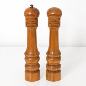 May include: Two tall, cylindrical wooden pepper and salt mills featuring a warm brown grain finish. Each mill has a rounded top, a series of horizontal grooves near the neck, and a flared base with two distinct rounded tiers for a stable grip.