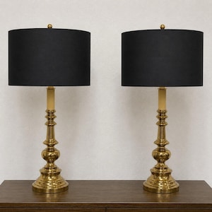 May include: A pair of table lamps featuring polished brass bases with a tiered, turned column design. Each lamp is topped with a cylindrical black fabric shade and a small brass finial at the center of the top edge of the shade.