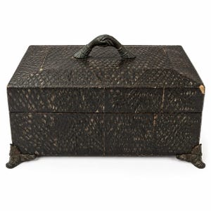 May include: A rectangular dark brown box with a textured, diamond-patterned surface. The lid features a handle shaped like two clasped hands, and the base is supported by four ornate, claw-like feet. The box has a hinged lid and a dark finish.