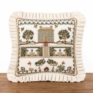 May include: A square off-white throw pillow featuring a ruffled border and intricate embroidery. The design depicts a central house, trees, birds, and garden elements in shades of green, brown, and red, creating a detailed, multi-layered scene.
