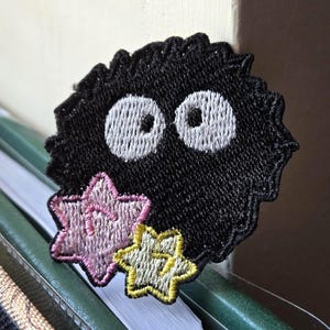 May include: Embroidered patch featuring a black, round creature with large white eyes and a spiky outline. Two star-shaped patches, one pink and one yellow, are below the creature. The patch is placed on top of a stack of folders.