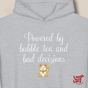 Può includere: Felpa con cappuccio grigia chiara con il testo bianco "Powered by bubble tea and bad decisions". Un gatto fortunato dei cartoni animati è sotto il testo. Il logo Laugh with Lucy si trova in basso a destra.