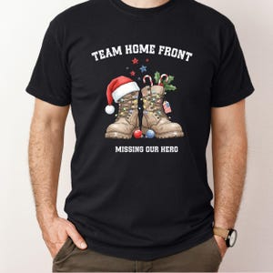 Peut inclure: T-shirt noir avec l'inscription "TEAM HOME FRONT" et "MISSING OUR HERO". Le graphisme représente des bottes de combat décorées de guirlandes lumineuses, d'un bonnet de Père Noël, de cannes en sucre et d'un drapeau américain.