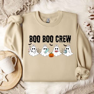 May include: A tan sweatshirt with the text "BOO BOO CREW" in black. Below the text are four cartoon ghosts wearing nurse hats, each holding a medical tool or pumpkin. Bats are scattered above the ghosts.