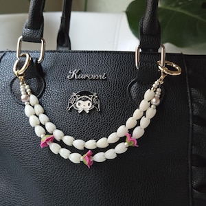May include: Black handbag with a decorative strap featuring white beads and pink floral accents. The bag has a silver "Kuromi" emblem and gold-colored hardware. The bag's handles are black.