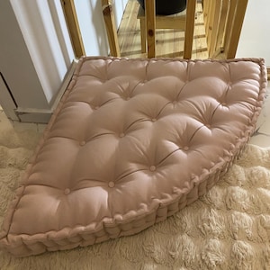 Puede incluir: Un cojín de suelo rosa rubor, con mechones y una esquina redondeada. El cojín tiene un diseño con botones y una construcción gruesa y acolchada. Se encuentra sobre una alfombra de pelo largo de color crema. Un elemento de decoración suave y acogedor.