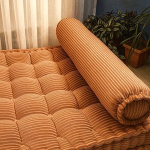 May include: A tufted floor cushion and a bolster pillow in a warm, orange-brown corduroy fabric. The cushion has a grid pattern, and the bolster is cylindrical. The items are placed near a window and plants.