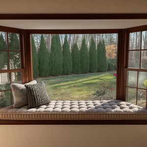 May include: A window seat with a tufted cushion and two decorative pillows. The window offers a view of a green lawn and tall evergreen trees. The window frame is made of dark wood.