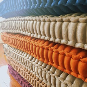 May include: A stack of colourful, tufted cushions in shades of blue, beige, orange, and purple. The cushions have a ribbed design and are stacked. They appear to be made of a soft, plush material. Each cushion is approximately 40 cm wide.