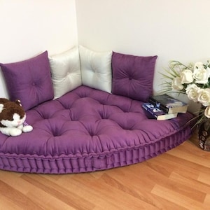 May include: A purple, semi-circular floor cushion with button detailing and matching cushions. A soft toy and books are placed on the cushion. A vase of white roses sits nearby, creating a reading corner.