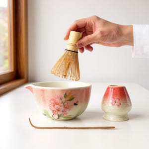 Op de afbeelding: Een matcha-theeset met een keramische kom met bloemmotieven, een bamboe klopper, een kleine keramische theekop en een bamboelepel. De kom en de beker hebben een roze en witte gradiënt. De klopper wordt boven de kom gehouden.