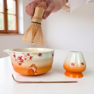 May include: A matcha tea set featuring a bamboo whisk, a ceramic bowl with a fox illustration, a small ceramic tea cup, and a bamboo scoop. The bowl and cup have a gradient orange and white color scheme with red leaf accents.