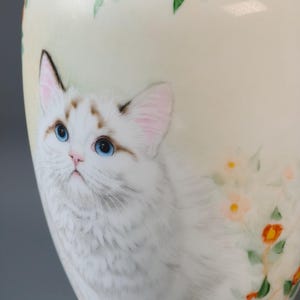 May include: A ceramic vase with a hand-painted design featuring a white cat with blue eyes and floral accents. The vase has a cream-colored background with orange and green flowers painted around the top.