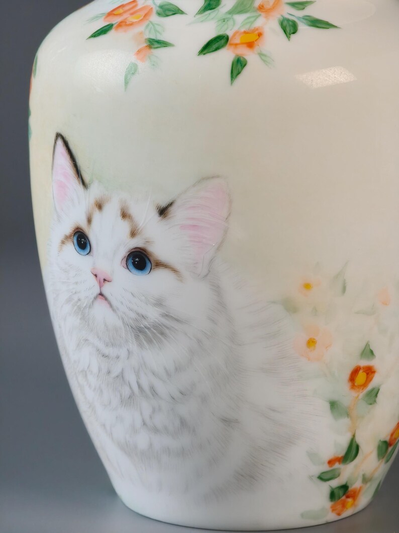 May include: A ceramic vase with a hand-painted design featuring a white cat with blue eyes and floral accents. The vase has a cream-colored background with orange and green flowers painted around the top.