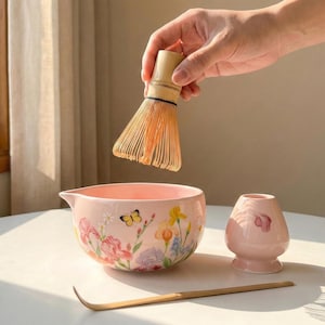 May include: A pink ceramic matcha bowl with floral designs, a bamboo whisk, a matcha scoop, and a pink ceramic whisk holder. The whisk is held above the bowl. The bowl has a spout.
