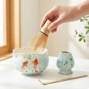 Puede incluir: Un juego de té matcha con un cuenco de cerámica azul claro con ilustraciones de peces koi, un batidor de bambú, un soporte para el batidor y una cuchara de bambú. El batidor se sostiene sobre el cuenco. El juego está sobre una superficie blanca.