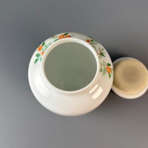 May include: A white ceramic jar with a lid, decorated with orange and green floral patterns. The jar has a wide opening and a round shape. The lid is off to the side, revealing the interior.