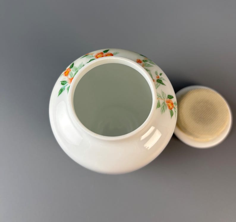 May include: A white ceramic jar with a lid, decorated with orange and green floral patterns. The jar has a wide opening and a round shape. The lid is off to the side, revealing the interior.