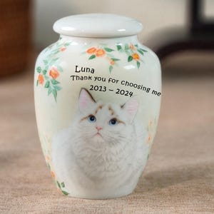 May include: A white ceramic pet urn with a lid, featuring a watercolor illustration of a cat with blue eyes. The urn is decorated with floral accents and includes the text "Luna, Thank you for choosing me 2013-2024."