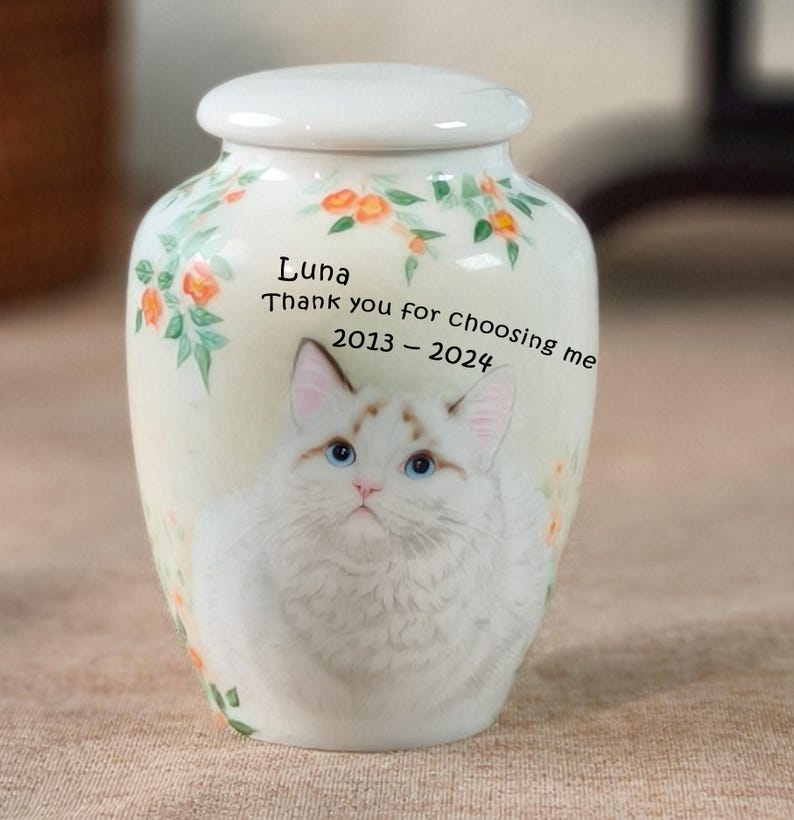 May include: A white ceramic pet urn with a lid, featuring a watercolor illustration of a cat with blue eyes. The urn is decorated with floral accents and includes the text "Luna, Thank you for choosing me 2013-2024."