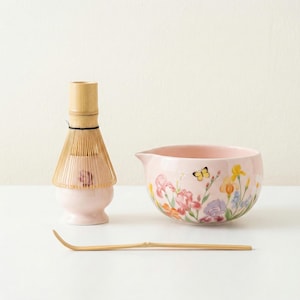 May include: A matcha tea set comprising a pink ceramic bowl with floral patterns, a bamboo whisk, and a bamboo scoop. The set is designed for preparing matcha tea.