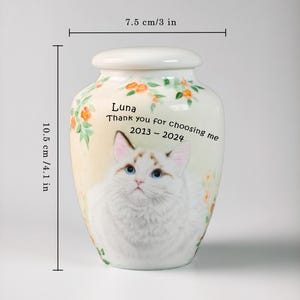 May include: A cream-colored ceramic urn, 4.1 inches tall and 3 inches wide, featuring a watercolor painting of a cat with blue eyes. The urn has floral accents and the text "Luna, Thank you for choosing me 2013-2024."