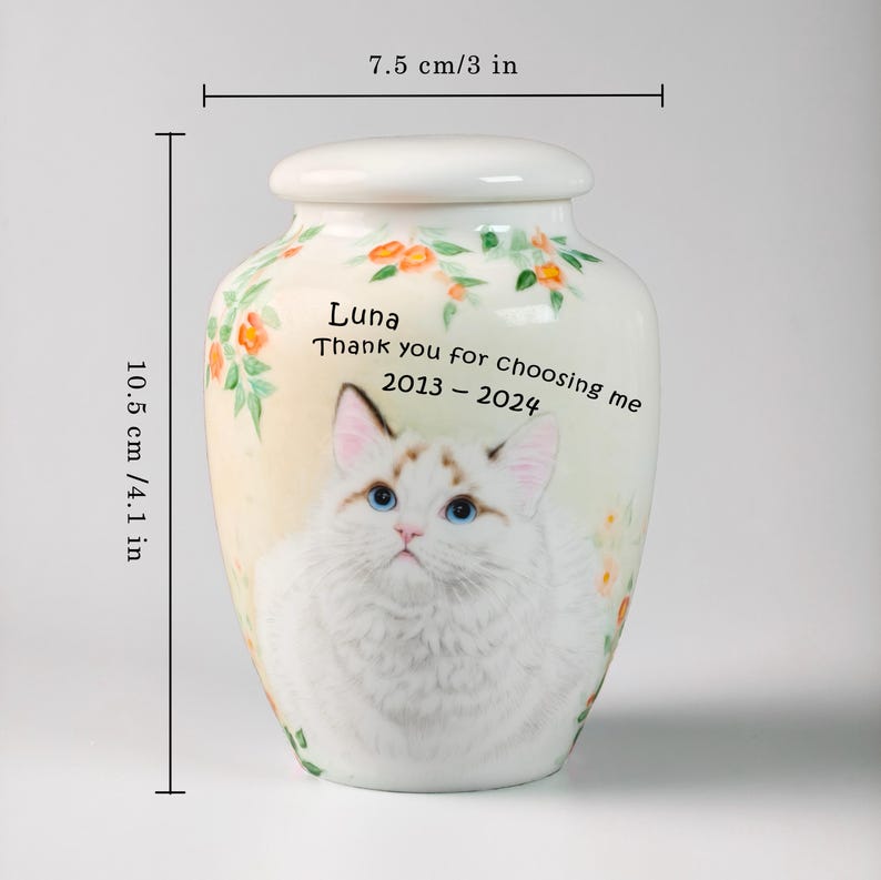May include: A cream-colored ceramic urn, 4.1 inches tall and 3 inches wide, featuring a watercolor painting of a cat with blue eyes. The urn has floral accents and the text "Luna, Thank you for choosing me 2013-2024."