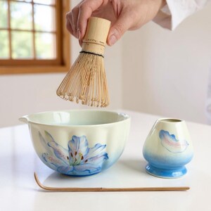 May include: A matcha tea set featuring a bamboo whisk, a ceramic bowl with a blue floral design, a small ceramic holder, and a bamboo scoop. The bowl and holder have a gradient blue base. The whisk is held above the bowl.