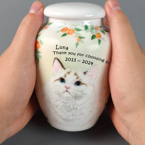 May include: A white ceramic urn with a lid, featuring a watercolor painting of a cat with blue eyes. The urn is decorated with orange flowers and green leaves. Text on the urn reads "Luna, Thank you for choosing me 2013-2024."