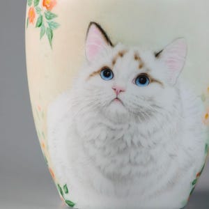 May include: A white ceramic vase featuring a detailed illustration of a fluffy white cat with striking blue eyes. The vase is adorned with floral accents in shades of orange and green, set against a soft, creamy background.