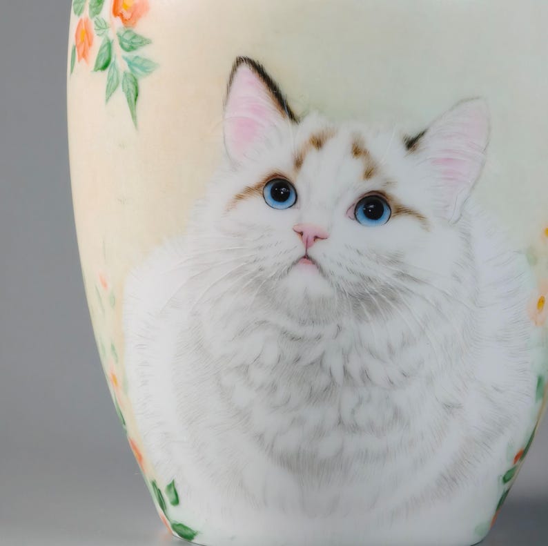 May include: A white ceramic vase featuring a detailed illustration of a fluffy white cat with striking blue eyes. The vase is adorned with floral accents in shades of orange and green, set against a soft, creamy background.