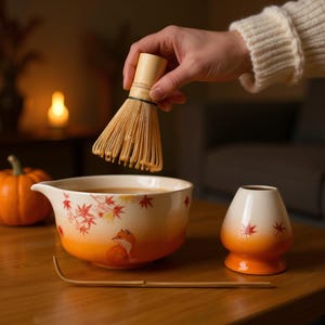 May include: A matcha tea set featuring a ceramic bowl with a fox illustration and maple leaf accents, a bamboo whisk, and a small ceramic tea bowl. The bowl has an orange gradient, and the whisk is held above the bowl.