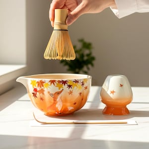 May include: A matcha tea set including a bamboo whisk, a ceramic bowl with a fox and autumn leaf design, a ceramic tea cup, and a bamboo tea spoon. The bowl and cup have orange and white gradients.