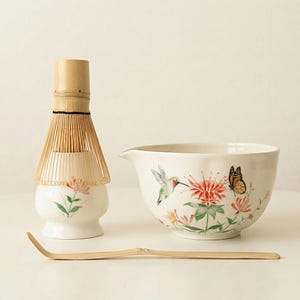 Puede incluir: Un juego de té matcha con un batidor de bambú, un cuenco de cerámica con un diseño floral y colibrí, y una cuchara de bambú. El cuenco tiene un pico vertedor. El batidor es marrón claro y el cuenco es blanco.