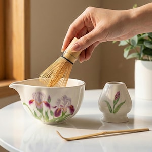 May include: A white ceramic matcha bowl with a purple iris floral design, a bamboo whisk, a small ceramic matcha holder, and a bamboo scoop. The whisk is held over the bowl.