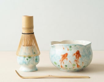 Könnte beinhalten: Ein Matcha-Teeset mit einem Bambusbesen, einer Keramikschale mit Koi-Karpfen-Design und einem Bambuslöffel. Die Schale hat einen hellblauen Hintergrund mit orangefarbenen Koi-Karpfen und grünen Seerosenblättern. Der Schneebesen ist aus hellbraunem Bambus.