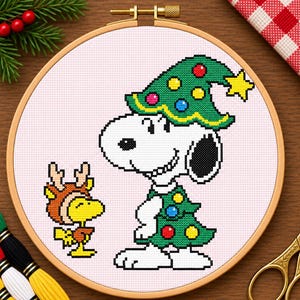 May include: A cross-stitch embroidery project in a wooden hoop featuring a white dog wearing a green Christmas tree costume with colourful ornaments and a matching hat, standing next to a small yellow bird wearing brown reindeer antlers.