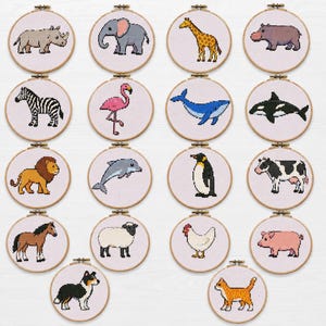Puede incluir: Una colección de diecisiete bastidores de bordado circulares de madera, cada uno con un diseño de punto de cruz de un animal diferente sobre tela blanca. Los animales incluyen un rinoceronte, elefante, jirafa, hipopótamo, cebra, flamenco, ballena, orca, león, delfín, pingüino, vaca, caballo, oveja, pollo, cerdo, perro y gato.