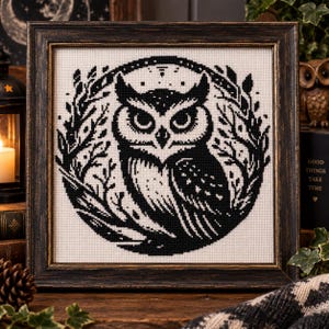 May include: A square framed cross-stitch embroidery featuring a black owl silhouette centered within a circular wreath of leaves and branches. The design is stitched in black thread on a white fabric background, enclosed in a dark distressed wooden frame.