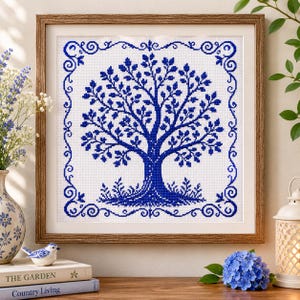 Può includere: Opera d'arte a punto croce incorniciata con un disegno di albero blu su sfondo bianco. L'albero è circondato da un bordo decorativo blu. L'opera è presentata in una cornice di legno.